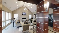 This single-floor space makes a strong architectural statement with its dramatically vaulted ceilings that draw the eye upward, creating an expansive sense of volume. The main living area is anchored by a sleek, linear fireplace set into a neutral-toned accent wall, above which a wall-mounted television is positioned for optimal viewing. Two contemporary chandeliers with an orb-style design hang from the ceiling, providing both ambient illumination and a decorative focal point, complemented by recessed lighting throughout.

The seating arrangement is thoughtfully composed, featuring multiple neutral-toned sofas and upholstered chairs arranged around area rugs, offering distinct conversation and lounging zones. A glass-top side table and a dark wood coffee table add functional elegance to the layout. Natural light filters in through multiple windows dressed with clean white roller shades.

A distinctive design element is the rich, dark wood-paneled column with horizontal metallic inlay strips that frames the entryway, adding a bold contrast to the otherwise light and airy color palette. The overall ambiance is polished and contemporary, balancing comfort with modern sophistication.