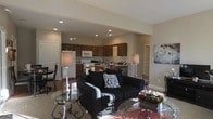 This single-floor residence offers an open-concept layout that integrates the living, dining, and kitchen areas into one cohesive and functional space. The living room serves as the heart of the home, furnished with a plush black sofa accented by bold zebra-print throw pillows, complemented by glass-topped coffee tables with decorative ironwork bases that add an elegant touch. Recessed lighting throughout the ceiling provides consistent, warm illumination, enhancing the neutral-toned walls that lend a clean and inviting atmosphere. A stylish floor lamp anchors the seating area, adding ambient warmth. The kitchen is equipped with dark wood cabinetry, a white refrigerator, built-in microwave, and a range, all arranged efficiently around ample counter space. A breakfast bar serves as a natural transition between the kitchen and living area, offering additional seating and a casual dining option. The open flow between spaces promotes a sense of spaciousness throughout. Decorative wall art adds a personal, curated touch to the living area. The overall ambiance is modern and comfortable, with thoughtful design details that make this single-floor layout both practical and welcoming for daily living.