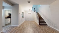 This two-story home offers a well-organized layout with a variety of thoughtfully designed spaces across both floors. The entryway makes a strong first impression with its warm wood-look flooring, neutral walls, and an elegant brass chandelier that anchors the open foyer. A carpeted staircase with a classic dark wood banister and white spindles rises gracefully to the upper level, while a pass-through window cutout adds architectural interest to the main level.

The galley-style kitchen is efficiently arranged, featuring white cabinetry with brushed nickel hardware, granite countertops, a subway tile backsplash accented with a mosaic tile border, a stainless steel sink, and matching appliances. Wood-look tile flooring runs throughout the kitchen, maintaining visual continuity with the rest of the home.

A standout feature of the living area is a full exposed brick accent wall, lending a warm, textural character to the space. Natural light filters in through a barred window, highlighting the rich tones of the hardwood-style flooring. A brick fireplace with a wood mantel adds to the room's charm.

The bathroom includes a wide vanity with white cabinetry, a light stone-tone countertop, undermount sink, and framed mirrors. Throughout, the home maintains a clean, neutral palette that provides a versatile and welcoming atmosphere across all areas.