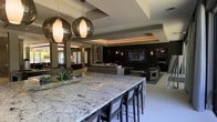 This single-story space showcases a refined open-plan layout that seamlessly connects the kitchen and living areas into one cohesive, contemporary environment. At the heart of the space is an expansive granite island with striking veining in cool grey and white tones, offering generous bar-style seating with dark upholstered chairs. Two large smoked-glass globe pendant lights hang overhead, providing both dramatic visual impact and warm ambient illumination.

The ceiling features elegant tray detailing with integrated cove lighting, adding architectural depth and a soft, layered glow throughout the space. The flooring is light-toned and continuous, reinforcing the open, airy feel of the layout.

The living area beyond is anchored by a large flat-screen television mounted on a dark feature wall flanked by built-in shelving and subtle wall sconces. Neutral, low-profile contemporary furniture defines the lounge zone without interrupting sightlines. A secondary entertainment or games area is visible in the background, further extending the functionality of the space. Natural light filters in from surrounding windows, enhancing the modern, welcoming ambiance throughout.
