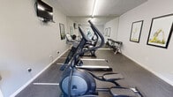 Located at 800 Valleyview Road in Pelham, Alabama, this fitness center offers a well-organized, single-room workout facility designed to support a range of exercise routines. The space features durable speckled rubber flooring throughout, providing both traction and cushioning underfoot. Overhead fluorescent strip lighting ensures the room is brightly and evenly illuminated.

The cardio section is well-appointed with multiple elliptical trainers, a treadmill, and a stationary recumbent bike, catering to varied cardiovascular training preferences. A full-length wall mirror runs along one side, visually expanding the space and allowing users to monitor their form. A free-weight rack holding an assortment of dumbbells is positioned along the mirrored wall for strength training.

Two wall-mounted flat-screen televisions are installed at separate points in the room, allowing users to stay entertained during workouts. A body weight scale is available for health tracking. Framed artwork depicting yoga poses adds a motivational aesthetic touch to the neutral gray walls, which feature a textured ceiling above.

A glass-paneled entry door allows natural light to filter into the space and offers views of the surrounding greenery outside. A wall-mounted water fountain and a sanitizing station near the entrance support hygiene and hydration. Fitness center rules are posted prominently near the door for user reference.