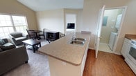 This single-floor space is designed with an open-concept layout that integrates the kitchen and living area seamlessly. The kitchen features a peninsula with a double stainless steel sink and laminate countertops, providing ample prep space and a natural dividing point between the kitchen and the adjoining living and dining areas. The flooring in the kitchen transitions from hardwood-style planks to carpet in the living and dining zones, adding textural variety throughout the space.

The dining area accommodates a small table and chairs, while the living area offers enough room for additional furnishings. Neutral beige walls and a textured ceiling keep the overall aesthetic clean and understated. A doorway from the main living area leads directly into a bedroom, which is visible in the background and appears to include a bed and basic furnishings. To the right of the kitchen, a frosted-glass door provides access to what appears to be a bathroom, offering privacy while allowing diffused light into the corridor. The overall layout is practical and efficiently organized, making good use of the available space on a single floor.