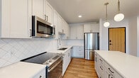 This thoughtfully designed two-bedroom, two-bathroom residence spans a comfortable single floor, offering a well-organized layout that includes a living room, kitchen, two bedrooms with dedicated closets, two bathrooms, and a hallway connecting the spaces.

The kitchen serves as a standout feature, outfitted with white shaker-style cabinetry with matte black hardware, stainless steel appliances including a range, built-in microwave, refrigerator, and dishwasher, and a diamond-patterned tile backsplash. White quartz countertops extend to a central island, which is illuminated by a trio of globe pendant lights, creating a functional and stylish focal point. Warm hardwood-style flooring runs throughout the kitchen and living areas.

The living room opens to a private balcony through a sliding glass door, allowing natural light to fill the space. Both bedrooms feature plush carpet flooring, large windows with exterior views, and wire-shelved closets providing organized storage. The bathrooms are finished with wood-look flooring, white tile surrounds, and clean, minimal fixtures. Overall, the unit presents a contemporary, light-filled aesthetic with cohesive finishes throughout.