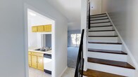 This two-story home presents a straightforward and functional layout designed for everyday living. The main level includes a kitchen equipped with warm honey-toned wood cabinetry, a double-basin sink, a dishwasher, and light countertops, providing ample storage and workspace. The kitchen opens visually to the adjacent living area, which features neutral carpeting and a sliding glass door that invites natural light and offers a view of the outdoors, creating a bright and airy atmosphere. The light, neutral color palette throughout the main level — with white walls and light tile flooring in the entryway — lends the space a clean and move-in-ready feel. A staircase with dark wood treads, white risers, and a contrasting dark handrail leads to the upper level, adding a classic design element to the interior. The transition between the kitchen and living spaces flows naturally, making the main floor feel cohesive and well-connected. Overall, this home combines practical features with a simple, neutral aesthetic suited to a variety of lifestyles.