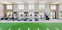 Located at 1450 Front Pl in North Port, Florida, this expansive fitness center offers a generously sized, single-floor exercise facility designed to support a wide range of workout routines. The space is defined by soaring ceilings that create an open, airy atmosphere, complemented by multiple tiers of large windows that flood the interior with abundant natural light and provide views of the surrounding Florida landscape.

A standout feature is the centrally positioned turf track with yard-line markings, ideal for agility drills, sled pushes, and functional training movements. Along one wall, a row of cardio machines — including multiple treadmills, ellipticals, and a stair climber — face wall-mounted flat-screen TVs and large glass doors that open to outdoor views. The strength training area is equally well-equipped, featuring power racks, a comprehensive dumbbell rack ranging across multiple weights, kettlebells, medicine balls, and various cable and plate-loaded machines. Large wall mirrors enhance the sense of space and support proper form during workouts.

The flooring transitions between turf and light-toned carpet, clearly delineating functional zones throughout the facility. Additional amenities include open shelving for storage, seating, and direct access points to the outdoors. The overall design balances a bright, modern aesthetic with practical, performance-focused equipment placement.