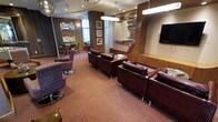 This single-floor lounge space exudes a sophisticated, contemporary ambiance with a warm and inviting atmosphere. The main seating area is furnished with deep burgundy leather armchairs and sofas arranged around low tables, offering comfortable gathering spots throughout the room. A patterned carpet in earthy rose and mauve tones adds texture and visual depth underfoot, complementing the rich color palette of the furnishings.

The décor is a standout feature, highlighted by an artistic wall installation of layered, staggered shelving elements that add dimension and a sculptural quality to the space. A large flat-screen television is mounted on the wall, flanked by a sleek wooden media console, making this area ideal for entertainment and viewing. Recessed lighting and modern pendant fixtures illuminate the space evenly, enhancing its polished feel.

An adjacent dining or meeting area is visible through a glass partition, furnished with round tables and chairs, offering a more intimate setting for smaller groups. The walls are adorned with framed artwork, further contributing to the curated, upscale interior design. Overall, the space strikes a balance between comfort and style, making it well-suited as a hospitality lounge, club room, or communal amenity space.