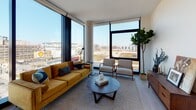 This single-floor residence is defined by its striking contemporary architecture, highlighted by expansive floor-to-ceiling windows framed in dark metal that wrap around the living area, flooding the interior with natural light and offering panoramic views of the surrounding urban cityscape. A structural concrete column adds an industrial accent to the open living space, where light hardwood flooring runs throughout.

The living area is generously proportioned, providing ample space for comfortable seating arrangements, including a bold mustard-yellow sofa and mid-century-style armchairs positioned to take full advantage of the sweeping city views.

The kitchen features white upper cabinetry, stainless steel appliances, and a large peninsula with a wood-panel base and white countertop, illuminated by a sleek linear pendant light. Upholstered bar stools complete the casual dining setup.

The bedroom continues the floor-to-ceiling window theme, framing bright city views and filling the room with daylight. A black-framed four-poster bed adds a modern design statement.

The bathroom is finished with large-format marble-look porcelain tile flooring, a wood-toned vanity, and clean white walls, creating a calm, spa-like atmosphere.