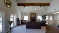 This single-floor residence makes an immediate impression with its soaring vaulted ceilings in the main living area, creating a grand sense of openness and volume. A prominent exposed wood beam runs the length of the ceiling, adding a warm, rustic architectural character that anchors the space. Track lighting mounted along the beam provides versatile illumination, complemented by a ceiling fan and recessed lighting throughout.

The living room serves as the heart of the home, centered around a classic full-height brick fireplace that adds both charm and a cozy focal point. Large windows flank the fireplace, inviting generous natural light into the room while providing views to the surrounding outdoors. A glass-paned door on one side further enhances the connection between the interior and exterior.

The space is finished with plush wall-to-wall carpeting that adds comfort underfoot and contributes to the room's warm, welcoming ambiance. The neutral wall tones and carefully considered architectural details — from the exposed timber to the brick masonry — work together to create a cohesive, inviting atmosphere. The overall layout is generous and flowing, making this home well-suited for both relaxed everyday living and entertaining guests.