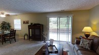 This single-floor space centers around a comfortable living room finished with wall-to-wall carpet and neutral olive-toned walls that create a warm, understated atmosphere. A textured popcorn ceiling adds a classic touch overhead, while a central air vent ensures year-round comfort. The room's standout feature is a large sliding glass door dressed with vertical blinds, which floods the space with natural light and provides direct views of a fenced backyard with lush greenery beyond. The door offers convenient access to the outdoor area, making the transition between indoor and outdoor living seamless. A dark wood hutch cabinet positioned near the entrance wall adds a traditional, functional element to the room's decor. The living area accommodates a coffee table and seating arrangement, providing a practical and inviting layout. Overall, the space conveys a cozy, functional character well-suited for everyday living, with the garden-facing glass door serving as a key architectural highlight that connects the interior to its natural surroundings.