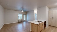 This single-floor unit offers a clean, contemporary design throughout. The open-concept layout connects the kitchen and living area seamlessly, creating a cohesive and functional living space. Light wood-tone laminate flooring runs throughout the main areas and bedroom, contributing to a warm, modern aesthetic.

The kitchen is well-appointed with light wood cabinetry extending to the ceiling for ample storage, white countertops, a peninsula with an undermount sink, and black appliances including a refrigerator, electric range, and over-the-range microwave. The peninsula provides a natural divide between the kitchen and living area.

The living area is bright and open, featuring a ceiling fan with a light fixture and a wall-mounted HVAC unit. A glass door and adjacent window invite natural light and provide access to an outdoor area with views of the surrounding building exterior.

The bedroom is a comfortable size with two windows that bring in natural light. The bathroom features a wood-tone vanity with white countertop, a mirror, a shower/tub combination, and distinctive patterned tile flooring that adds visual interest to the neutral space.