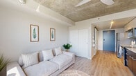 Located at 404 Strander Blvd in Tukwila, WA, this single-floor studio unit offers a thoughtfully designed open-concept layout that maximizes every inch of available space. The interior is defined by striking exposed concrete ceilings, which lend an industrial-modern character throughout, contrasted by clean white walls and warm light wood flooring that create a bright, cohesive aesthetic.

The kitchen is efficiently arranged along one wall, featuring dark cabinetry with a contrasting white countertop and upper cabinets, stainless steel appliances including a refrigerator and range, and a sleek single-basin sink. A sliding barn door adds a stylish functional detail near the entry. A small dining area with a round white table sits adjacent, making efficient use of the open floor plan.

The living area is airy and bathed in natural light from a large window that frames an elevated outdoor view of the surrounding Tukwila landscape. The space comfortably accommodates a full-sized sofa and area rug, with the exposed concrete ceiling continuing to reinforce the unit's contemporary industrial tone.

The bathroom is clean and functional, finished with the same light wood-style flooring, and includes a full shower-tub combination, toilet, and ample wall space. The overall ambiance of the unit is modern, minimal, and efficiently organized.