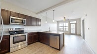 This thoughtfully designed one-bedroom, one-bathroom unit offers an efficient and functional layout across a single floor. The open-concept kitchen and living area serves as the heart of the unit, featuring rich dark-espresso cabinetry, granite countertops, a peninsula island with pendant lighting overhead, and a full suite of stainless steel appliances including a range, microwave, dishwasher, and refrigerator. Light hardwood-style flooring runs throughout the main living areas, complementing the neutral wall tones and creating a warm, cohesive aesthetic. A ceiling fan anchors the living space, which is bathed in natural light from large windows and a glass-paneled door leading to a private balcony with outdoor views. The bedroom is carpeted for comfort, offering a ceiling fan and a generous window with additional natural light. It connects to a spacious walk-in closet equipped with wire shelving for ample storage. The bathroom features hardwood-style flooring consistent with the main living areas, dark vanity cabinetry with granite countertops, and direct hallway access to the closet. The unit's clean, modern design and practical layout make it a highly functional living space.