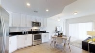 This single-floor residence offers two bedrooms and two bathrooms within a thoughtfully designed open-concept floor plan. The kitchen serves as a focal point of the living space, outfitted with crisp white shaker-style cabinetry, dark contrasting countertops, and stainless steel appliances including an electric range and over-the-range microwave. Recessed lighting illuminates the kitchen area, while a decorative pendant light marks the transition into the dining space.

The dining area accommodates a modest table and seating, flowing seamlessly into the living area, which benefits from a large sliding door or window treatment that invites natural light into the space. Light-toned hardwood-style flooring runs throughout, reinforcing the clean, cohesive aesthetic. The tray ceiling detail adds a subtle architectural element, lending a sense of dimension to the otherwise streamlined interior.

Overall, the space maintains a bright, airy ambiance with a neutral white-and-light palette, modern fixtures, and an efficient layout that maximizes functionality across the open living, dining, and kitchen zones.