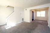 Located at 3817 George Washington Hwy in Portsmouth, Virginia, this two-floor home offers a practical and comfortable layout designed for everyday living. The main level features a spacious living area with plush wall-to-wall carpeting, neutral-toned walls, and a window that brings in natural light. Adjacent to the living space is a dedicated dining area, distinguished by a classic multi-bulb chandelier overhead and direct access to adjoining rooms. The kitchen is equipped with warm oak cabinetry — both upper and lower — providing generous storage, along with a double stainless steel sink, light countertops, and a black electric range with overhead ventilation. The main level also includes a door leading to the exterior. A staircase connects the main level to the upper floor, where the bedrooms are located. Each bedroom features soft carpeting, neutral walls, and individual closets with built-in shelving, offering practical storage solutions. The bathroom includes a full bathtub and shower combination with clean white surrounds and a window allowing natural light. Throughout the home, consistent use of neutral tones, white trim, and carpet creates a cohesive and move-in ready atmosphere suited for a variety of lifestyles.