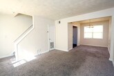 Located at 3817 George Washington Hwy in Portsmouth, Virginia, this two-floor home offers a practical and comfortable layout designed for everyday living. The main level features a spacious living area with plush wall-to-wall carpeting, neutral-toned walls, and a window that brings in natural light. Adjacent to the living space is a dedicated dining area, distinguished by a classic multi-bulb chandelier overhead and direct access to adjoining rooms. The kitchen is equipped with warm oak cabinetry — both upper and lower — providing generous storage, along with a double stainless steel sink, light countertops, and a black electric range with overhead ventilation. The main level also includes a door leading to the exterior. A staircase connects the main level to the upper floor, where the bedrooms are located. Each bedroom features soft carpeting, neutral walls, and individual closets with built-in shelving, offering practical storage solutions. The bathroom includes a full bathtub and shower combination with clean white surrounds and a window allowing natural light. Throughout the home, consistent use of neutral tones, white trim, and carpet creates a cohesive and move-in ready atmosphere suited for a variety of lifestyles.
