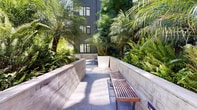 This outdoor courtyard offers a serene urban retreat set within the footprint of a contemporary multi-story building. The space is defined by its clean, modern design, featuring large-format porcelain or stone pavers in a soft gray tone that extend across the entire ground level, lending the area a polished, cohesive aesthetic.

Raised stone-clad planters serve as both structural dividers and garden beds, overflowing with a rich variety of tropical and subtropical plantings including towering palm trees, dense ferns, and lush shrubs. These plantings create a verdant, almost enclosed atmosphere, softening the angular lines of the surrounding building facade and providing a strong sense of privacy and natural immersion.

Seating is provided through a series of slatted wood-and-metal benches positioned throughout the courtyard, offering comfortable resting spots that complement the modern material palette. Built-in step lighting integrated into the planter walls adds a warm, functional glow and enhances the ambiance during evening hours.

The surrounding building features a contemporary exterior with dark-framed windows, smooth panel cladding in deep gray-green tones, and visible balconies. Abundant natural light filters into the courtyard, casting dynamic shadows across the pavers and highlighting the textures of the stone and foliage. Overall, the space strikes a balance between structured urban design and organic natural beauty.