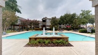 The Main Pool area is a well-appointed outdoor amenity space designed for relaxation and recreation. At the heart of the space sits a large, resort-style swimming pool with clear turquoise water, complemented by a striking stone-accented decorative fountain feature at its center, with water jets that create an inviting focal point. The pool deck is finished with textured concrete and stone tile surrounds, providing ample space for lounge seating along the perimeter.

A pergola-covered outdoor lounge area adjacent to the pool offers shaded seating with bar-height chairs and tables, providing a comfortable gathering spot. Red market umbrellas add a pop of color and additional shade to the patio seating areas. A secondary covered pavilion with arched columns is also present, enhancing the architectural character of the space.

Access to the pool area is controlled through an ornate black wrought iron gate set beneath a stuccoed archway entry structure, adding a sense of security and elegance. Well-maintained landscaping, including manicured shrubs, flowering crape myrtles, mature trees, and mulched garden beds, frames the entire space, creating a lush, park-like atmosphere. The surrounding multi-story residential buildings provide a scenic backdrop throughout.