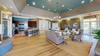Located at 56 Bent Branch Loop in Clayton, NC, this single-story community clubhouse offers a thoughtfully designed and generously sized gathering space for residents. The welcoming entry foyer sets the tone with soaring ceilings, soft sage-blue walls, crisp white millwork, and light hardwood flooring that flows through the hallways connecting each area of the facility.

The heart of the clubhouse is a expansive open-plan lounge and social area featuring a tray ceiling accented in a bold teal hue and anchored by an elegant chandelier. A full kitchen with rich wood cabinetry, granite countertops, and a breakfast bar serves the space seamlessly. Multiple seating arrangements create an inviting, flexible environment ideal for both large gatherings and quiet relaxation.

The fitness center is a standout amenity, divided into distinct zones for cardio and strength training. It is outfitted with treadmills, ellipticals, a stationary bike, a multi-station cable machine, and a full dumbbell rack. Floor-to-ceiling mirrors, rubber flooring, and abundant natural light from multiple windows make this a fully functional workout facility.

A dedicated business office provides dual workstations with built-in granite-topped desks and computer stations beneath a branded logo display. Additional rooms include cozy seating nooks with warm wood-paneled ceilings, completing this well-appointed community hub.