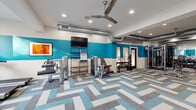 This fitness center at 740 Sidney Marcus Blvd NE in Atlanta, Georgia, offers a comprehensive workout environment designed to accommodate a wide range of exercise needs. The space is defined by a bold two-tone color scheme, with vibrant teal accent walls contrasting against crisp white upper walls, creating an energetic and motivating atmosphere throughout. The flooring features a striking herringbone-patterned carpet tile in shades of teal, gray, and beige, adding visual interest underfoot while providing a comfortable surface for workouts. Exposed industrial-style ductwork runs along the ceiling, complemented by recessed lighting and ceiling fans that ensure the space remains bright and well-ventilated. Large windows along one wall allow natural light to flow in, offering views of the surrounding community. The cardio zone is equipped with multiple treadmills, ellipticals, and a stationary bike, while the strength training area features a comprehensive dumbbell rack, cable machines, weight benches, and functional training equipment including kettlebells and medicine balls of varying weights. Wall-to-wall mirrors in the strength section enhance the sense of space and allow users to monitor their form. A wall-mounted flat-screen TV adds convenience for those looking to stay entertained during their workout sessions.