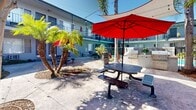 This inviting outdoor courtyard at 2353 Portland St in Los Angeles, CA offers a well-appointed communal gathering space nestled within a multi-story residential building. The courtyard is framed by the building's blue-toned exterior and wraparound balconies, creating an enclosed yet open-air atmosphere that captures the Southern California sunshine throughout the day.

At the heart of the space, mature palm trees and a large shade tree provide natural canopy, complemented by lush tropical plantings and landscaped gravel beds that add texture and greenery. Multiple metal picnic tables — each shaded by bold red umbrellas — offer comfortable seating for outdoor dining and socializing.

A standout feature is the built-in outdoor kitchen station, equipped with two full-sized stainless steel grills set into a stone countertop, perfect for communal barbecues. Overhead string lights are strung across the courtyard, creating a warm and welcoming ambiance for evening gatherings.

The courtyard also provides access to a pool area, visible through wrought iron fencing, adding a resort-like quality to the space. The combination of functional amenities, abundant natural light, and thoughtful landscaping makes this courtyard a lively and practical outdoor retreat in the heart of Los Angeles.