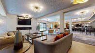 Located at 11 Maple Ave in Bay Shore, NY, this thoughtfully designed single-floor communal space blends contemporary elegance with comfortable, inviting interiors. The main lounge area makes an immediate impression with its eye-catching geometric patterned ceiling, statement Sputnik-style chandeliers, and a sleek marble-surround electric fireplace flanked by a wall-mounted flat-screen TV. Plush seating in warm neutral tones with bold rust-colored accent pillows adds a welcoming depth to the space. Herringbone-patterned hardwood flooring flows through connecting areas, complemented by richly textured area rugs in abstract designs. A café-style bar area features a white high-top communal table with modern counter seating, pendant lighting with warm gold accents, a distinctive wave-patterned textured feature wall, and built-in beverage and coffee stations. An adjacent game room showcases exposed stone accent walls, large windows that draw in natural light, and ample open floor space anchored by a foosball table. Throughout the space, curated artwork, tall indoor plants, and layered lighting create a sophisticated yet relaxed atmosphere, making this an exceptional gathering destination for its community.