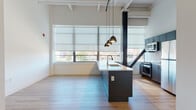 Located at 15 Van Dyke Ave in Hartford, Connecticut, this single-floor unit blends contemporary design with industrial-loft character. The open-concept living and kitchen area serves as the heart of the space, anchored by a sleek dark navy island with a crisp white countertop and an undermount sink. Pendant lighting hangs overhead, while the kitchen is equipped with stainless steel appliances, a built-in microwave, and a subway tile backsplash framed by matching dark cabinetry. Exposed ceiling beams, structural columns, and open ductwork add an authentic industrial aesthetic throughout. Large windows dressed with roller shades flood the interior with natural light and offer sweeping views of the Hartford skyline and surrounding urban landscape. Light-toned wood-style flooring flows consistently throughout, complementing the bright, neutral wall palette. The bedroom features soaring ceilings, rich wood-toned doors, and a built-in closet, with a large window that frames striking city views. The bathroom is thoughtfully designed for accessibility, complete with grab bars, a soaking tub, and tile flooring. The layout is generously proportioned, offering a comfortable and functional living environment with a distinctly modern urban character.