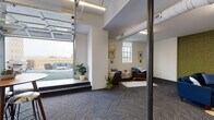 This single-floor space showcases a thoughtfully designed open-plan layout that blends industrial character with contemporary comfort. Exposed ductwork runs along the ceilings, complementing the textured green-painted brick accent wall that serves as a striking focal point throughout the main lounge area. Dark, patterned carpet tiles ground the space and extend throughout, providing a cohesive visual flow between zones.

The lounge area is anchored by deep navy blue sofas arranged around a round white coffee table, all facing a wall-mounted flat-screen television on the green brick wall. Recessed and linear fluorescent lighting illuminates the space evenly, while multiple windows allow natural light to filter in from multiple directions.

A separate dining and meeting area features a glass-top table surrounded by mid-century modern chairs, a built-in white cabinetry unit with a subway tile backsplash, a vibrant olive-green accent wall, and a high-top table with bar stools — ideal for casual gatherings or collaborative work sessions.

A standout feature is the large glass-panel garage door that opens directly onto a rooftop terrace, equipped with outdoor seating and a grill, seamlessly connecting the interior to the outdoors. The overall ambiance is modern, functional, and inviting.