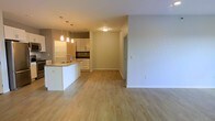 This single-floor space showcases a clean, modern design centered around an open-concept layout that seamlessly connects the kitchen and living areas. The kitchen is well-appointed with white cabinetry, a dark tile backsplash, and a peninsula island featuring a built-in sink — an ideal setup for both everyday cooking and casual entertaining. Pendant lighting hangs above the island, adding warm, focused illumination that complements the overall brightness of the space. Light-toned hardwood flooring runs throughout, lending a sense of warmth and continuity across the open floor plan. The living area is spacious and versatile, offering ample room for a variety of furniture arrangements. Neutral white walls provide a clean, contemporary backdrop that allows for easy personalization. A hallway leads to additional areas of the unit, suggesting a practical and well-organized floor plan. Ceiling-mounted lighting fixtures ensure consistent illumination throughout. Overall, the space conveys a fresh, move-in-ready aesthetic with a balance of functionality and modern style, making it well-suited for a range of occupants.