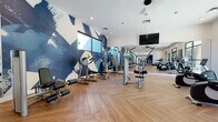 Located at 3400 E Southern Ave in Phoenix, AZ, this fitness center offers a generously sized, single-floor open layout designed to accommodate a full range of workout activities. The space is defined by eye-catching herringbone-patterned hardwood flooring that runs throughout, lending a modern and polished aesthetic to the environment.

A bold, large-scale blue abstract mural serves as a dramatic focal point on one wall, adding a vibrant, energetic character to the space. Full-length mirrors line portions of the walls, amplifying the sense of openness and allowing users to monitor their form during workouts. Wall-mounted flat-screen televisions are positioned for visibility across the floor.

The cardio section is well-appointed with treadmills, ellipticals, stationary bikes, and a rowing machine, all positioned near floor-to-ceiling windows and glass doors that frame outdoor views of the surrounding landscaped grounds and palm trees. Natural light flows generously into this area, creating a bright and motivating atmosphere.

The strength training section features a multi-station cable and weight machine system, adjustable benches, free weights, and medicine balls. Ceiling fans and recessed lighting provide comfortable, well-distributed illumination throughout. The glass door entry and large windows maintain a strong visual connection to the outdoor environment, enhancing the overall experience of this modern fitness facility.
