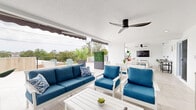 Located at 527 N Garden Ave in Clearwater, Florida, this thoughtfully designed outdoor space offers a seamless blend of covered and open-air living areas atop a single-story structure. The rooftop terrace is finished with large-format light-toned porcelain tiles throughout, lending the space a clean, modern aesthetic that complements the coastal Florida setting.

The covered section functions as a fully equipped outdoor living room, featuring white-framed furniture with vibrant blue cushioned seating, a lounge sofa, matching accent chairs, and a dining table with ample seating. A wall-mounted flat-screen TV and dual ceiling fans with sleek black blades make this area ideal for entertaining in comfort. A grill station rounds out the amenities, adding outdoor cooking functionality.

Retractable awnings extend over portions of the covered area, providing flexible shade and weather protection. Beyond the covered zone, the open-air terrace offers additional lounge seating, including Adirondack chairs, along with decorative tall planters featuring tropical foliage and palms that enhance the lush, resort-like atmosphere.

The perimeter is lined with modern cable-rail fencing, offering unobstructed panoramic views of the surrounding tree canopy and Clearwater skyline. A warm wood-panel accent wall adds natural texture to one corner of the open deck. The overall ambiance is bright, airy, and inviting — a true outdoor retreat designed for year-round Florida living.