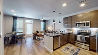 This single-floor residence features a well-designed open-concept layout that seamlessly connects the kitchen, dining, and living areas. The kitchen is outfitted with dark espresso cabinetry reaching toward the ceiling, dark granite countertops, a white subway tile backsplash, and a full suite of stainless steel appliances including a refrigerator, range, microwave, and dishwasher. A central island with seating provides additional prep space and casual dining. Pendant lighting accents the kitchen, while recessed lighting and ceiling fans serve the living area, which opens to an exterior door allowing natural light to fill the space. Light wood-style flooring runs throughout the main living areas, creating a cohesive and clean aesthetic. The dining area features large wall art and window access, contributing to a bright, functional space. Two bedrooms are finished with plush carpeting, multiple windows with blinds and curtains, and ceiling fans. The bathroom is accessed via a hallway that also connects to a walk-in closet, and features a tub-shower combination with a curved rod and wood-style flooring. Neutral gray walls throughout the unit provide a versatile, contemporary backdrop.