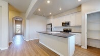 This single-floor home offers a clean, contemporary design with a thoughtful open layout that connects the main living areas seamlessly. The kitchen serves as the centerpiece of the home, featuring crisp white cabinetry, a classic subway tile backsplash, dark countertops, and a large center island complete with an integrated sink. Stainless steel appliances — including a refrigerator, range, and microwave — add a modern, functional touch. A built-in shelving nook adjacent to the kitchen provides additional storage or workspace. Light wood-tone flooring runs throughout the main living and entryway areas, creating a warm and cohesive aesthetic, while recessed lighting illuminates the space efficiently. The entryway welcomes natural light through a front door with glass panel insets and a large window fitted with white blinds. The bedrooms are carpeted in neutral tones and receive natural light through generously sized windows. A full bathroom includes a tub-shower combination with clean white tile surround and wood-look flooring. Throughout the home, arched architectural details and neutral wall tones contribute to an airy, inviting atmosphere well-suited for comfortable everyday living.