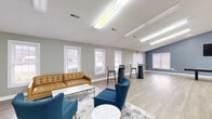 This well-appointed single-floor space offers a generous and flexible layout designed to accommodate a variety of gatherings and activities. The centerpiece is a expansive open hall with soaring vaulted ceilings, light wood-look flooring, and abundant overhead fluorescent lighting. Neutral gray walls and crisp white trim create a clean, modern aesthetic throughout. The hall is furnished with multiple round dining tables and upholstered chairs, making it ideal for meetings, events, or communal dining. French doors with glass panes connect sections of the space, allowing natural light to flow freely and providing visual openness. A dedicated lounge area features a tufted tan leather sofa, navy blue accent chairs, and stylish high-top tables with bar stools arranged along a row of white-trimmed windows. A separate game room includes a shuffleboard table and a wall-mounted television beneath a sloped ceiling, offering a casual recreational retreat. The fully equipped kitchen provides warm oak cabinetry, white countertops, a full-size refrigerator, electric range, dishwasher, and a movable island, making catering and food service convenient. Together, these distinct zones create a functional and inviting multi-purpose facility suited for community use, social events, or organizational gatherings.