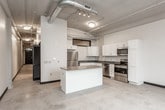 This single-floor unit exhibits a distinctive industrial aesthetic, with exposed ductwork running along the ceilings and polished concrete floors throughout. The open layout creates a sense of spaciousness, with the overall footprint extending in a long, rectangular configuration as revealed by the overhead dollhouse view.

The kitchen is a functional centerpiece of the unit, outfitted with white shaker-style cabinetry, granite countertops, and a full suite of stainless steel appliances, including a refrigerator, range, built-in microwave, and dishwasher. A kitchen island with a matching granite surface provides additional prep and storage space. Overhead, exposed industrial ductwork and a large ventilation hood add to the loft-like character of the space.

The bedroom area features painted white brick walls, lending a bright yet textured appearance. A large arched window allows natural light to fill the room while offering outdoor views, and a baseboard heating unit runs along the exterior wall. The bedroom is accessed through a bold black interior door, which contrasts cleanly against the light-toned walls and concrete floors.

The bathroom is cleanly appointed with a white vanity, granite countertop, undermount sink, recessed medicine cabinet mirror, and chrome fixtures, providing a tidy and practical finish to the space.