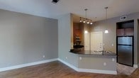 This single-floor space features an open-concept design that seamlessly connects the living area and kitchen. The living space is finished with warm hardwood-style flooring and neutral gray walls accented by crisp white baseboards, creating a clean and contemporary aesthetic.

The kitchen is defined by a curved peninsula counter finished in dark granite, providing both workspace and a natural division between the cooking and living areas. Overhead, a combination of track lighting and two pendant lights illuminate the kitchen, adding a modern touch. Warm-toned wooden cabinetry lines the kitchen walls, complemented by stainless steel appliances including a microwave and range. A stainless steel sink is integrated into the peninsula countertop.

The entry door, finished in white with a deadbolt, is visible from the main living area, reinforcing the open sightlines throughout the space. The neutral color palette, consistent flooring, and layered lighting contribute to an overall ambiance that feels functional, modern, and inviting.