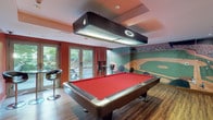 The Billiards Lounge is a well-appointed recreational space designed for social gaming and relaxation. The centerpiece of the room is a full-size pool table with bold red felt, illuminated by a large overhead pendant light fixture that ensures optimal visibility during play. Warm hardwood flooring runs throughout, complementing the room's energetic color palette of deep reds and charcoal tones.

A striking floor-to-ceiling baseball stadium mural wraps around a significant portion of the walls, creating an immersive, sports-bar atmosphere. On the opposite end of the room, a shuffleboard table sits beneath a wall-mounted flat-screen television, alongside a framed sports print, making it an ideal spot for friendly competition. A built-in fireplace with a stone surround anchors the back wall, adding warmth and ambiance to the space.

Stylish bar-height tables and sleek swivel stools are arranged throughout the lounge, providing comfortable seating for guests. Large glass doors along one wall open directly to an outdoor patio area, allowing ample natural light to fill the space and offering a seamless indoor-outdoor connection. Recessed ceiling lighting supplements the natural light, ensuring the room is well-lit at any hour. The Billiards Lounge delivers a dynamic, inviting environment suited for leisure and social gatherings alike.