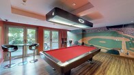 The Billiards Lounge is a well-appointed recreational space designed for social gaming and relaxation. The centerpiece of the room is a full-size pool table with bold red felt, illuminated by a large overhead pendant light fixture that ensures optimal visibility during play. Warm hardwood flooring runs throughout, complementing the room's energetic color palette of deep reds and charcoal tones.

A striking floor-to-ceiling baseball stadium mural wraps around a significant portion of the walls, creating an immersive, sports-bar atmosphere. On the opposite end of the room, a shuffleboard table sits beneath a wall-mounted flat-screen television, alongside a framed sports print, making it an ideal spot for friendly competition. A built-in fireplace with a stone surround anchors the back wall, adding warmth and ambiance to the space.

Stylish bar-height tables and sleek swivel stools are arranged throughout the lounge, providing comfortable seating for guests. Large glass doors along one wall open directly to an outdoor patio area, allowing ample natural light to fill the space and offering a seamless indoor-outdoor connection. Recessed ceiling lighting supplements the natural light, ensuring the room is well-lit at any hour. The Billiards Lounge delivers a dynamic, inviting environment suited for leisure and social gatherings alike.