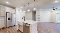 This thoughtfully designed 2-bedroom, 2-bathroom home in Austin, TX offers a clean, modern aesthetic throughout its single-floor layout. The open-concept kitchen and living area serves as the heart of the home, anchored by a striking central island with quartz countertops, a farmhouse-style sink with a matte black faucet, and a stainless steel dishwasher. White shaker cabinetry lines the kitchen walls alongside a full suite of stainless steel appliances, including a range, microwave, and refrigerator. Three globe pendant lights with brushed gold fixtures hang above the island, adding a warm, contemporary touch. Wood-look flooring flows seamlessly throughout the living areas and bedrooms, complementing the bright, neutral color palette. Built-in shelving in the living area provides functional display and storage space. The primary bedroom is generously sized and connects to a well-appointed walk-in closet with custom shelving and hanging rods. Both bathrooms feature marble-look tile surround tub-shower combinations, light cabinetry with matte black hardware, and large-format floor tiles. The second bedroom is bright and airy, benefiting from natural light through large windows and a ceiling fan overhead.