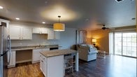 This single-floor space is designed with an open-concept layout that promotes a seamless flow between the kitchen and living areas. The kitchen is a standout feature, showcasing white cabinetry, granite countertops, and a central island with bar seating — ideal for casual dining or entertaining. A stylish drum pendant light hangs above the island, adding a warm, inviting glow to the space. The kitchen also includes a sink along the perimeter counter and built-in appliances for a clean, functional aesthetic. Dark hardwood-style flooring runs throughout, grounding the space with a rich, cohesive look that contrasts beautifully with the light-toned walls and white cabinetry. The living area transitions naturally from the kitchen, featuring a comfortable sofa, a ceiling fan for added comfort, and warm ambient lighting from a table lamp. A decorative wall piece with a rustic wood-panel design adds character and texture to the living space. Recessed lighting and neutral wall tones contribute to the overall bright and airy atmosphere. The thoughtful layout and cohesive design elements make this space both practical and aesthetically appealing for everyday living.