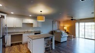 This single-floor space is designed with an open-concept layout that promotes a seamless flow between the kitchen and living areas. The kitchen is a standout feature, showcasing white cabinetry, granite countertops, and a central island with bar seating — ideal for casual dining or entertaining. A stylish drum pendant light hangs above the island, adding a warm, inviting glow to the space. The kitchen also includes a sink along the perimeter counter and built-in appliances for a clean, functional aesthetic. Dark hardwood-style flooring runs throughout, grounding the space with a rich, cohesive look that contrasts beautifully with the light-toned walls and white cabinetry. The living area transitions naturally from the kitchen, featuring a comfortable sofa, a ceiling fan for added comfort, and warm ambient lighting from a table lamp. A decorative wall piece with a rustic wood-panel design adds character and texture to the living space. Recessed lighting and neutral wall tones contribute to the overall bright and airy atmosphere. The thoughtful layout and cohesive design elements make this space both practical and aesthetically appealing for everyday living.