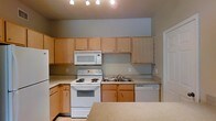 The kitchen is designed with practicality in mind, showcasing a clean and functional layout that maximizes both storage and workspace. Light wood cabinetry lines the walls, extending from lower cabinets to upper units that reach toward the ceiling, providing generous storage throughout. The cabinetry features simple hardware, contributing to a streamlined, understated aesthetic.

White appliances are integrated throughout, including an electric coil range with oven, an over-the-range microwave, a dishwasher, and a refrigerator positioned along the far wall. The double-basin stainless steel sink is centrally located along the counter run, offering a practical focal point for the workspace. Light-toned laminate countertops extend across the full length of the kitchen, providing ample prep area.

A peninsula or breakfast bar is partially visible in the foreground, offering an additional surface that could serve as a casual dining or prep space. Track lighting overhead ensures the kitchen is well-lit. The walls are painted in a neutral gray tone, complementing the light wood cabinetry and creating a cohesive, contemporary feel throughout the space.