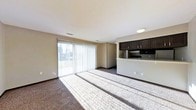 This single-floor space features an open-concept layout that seamlessly connects the living area to the kitchen. The living room is finished with wall-to-wall carpeting and is bathed in natural light from a large sliding glass door dressed with vertical blinds, offering a view of the surrounding outdoor greenery. The light filtering through the blinds casts distinct lines across the floor, enhancing the sense of openness in the room.

The kitchen is positioned at the rear of the open space, defined by a raised breakfast bar that serves as a natural divider between the two areas. Dark espresso-toned cabinetry lines the upper wall, providing a contrasting accent against the neutral beige walls and white ceiling. A built-in microwave or range hood is visible beneath the upper cabinets, indicating a functional cooking setup. Recessed and flush-mount ceiling lighting provides additional illumination throughout.

The neutral color palette of beige walls, white ceilings, and dark trim creates a clean, cohesive aesthetic. The layout is practical and straightforward, making efficient use of the available space while maintaining an airy, open feel throughout.
