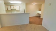 This single-floor space offers a functional and efficiently designed layout that maximizes every corner of the unit. The living area is finished with warm beige carpeting, creating a comfortable and inviting atmosphere that flows seamlessly into the adjoining spaces.

The kitchen is positioned behind a half-wall peninsula, which serves as both a practical workspace and a visual divider between the cooking area and the main living space. White cabinetry lines the kitchen walls, offering ample storage, while a full-size refrigerator and additional appliances complete the functional setup. Flush-mounted lighting above the kitchen workspace ensures the area is well-lit.

Adjacent to the kitchen, a dedicated nook area transitions to warm wood-tone laminate flooring, setting it apart from the carpeted living space. This nook features built-in open shelving and a built-in desk surface, making it ideal for use as a home office, study area, or organizational space.

The interior is finished with clean white walls and white baseboards throughout, giving the space a bright, neutral aesthetic that is easy to personalize. Ceiling-mounted light fixtures provide consistent illumination across the unit. The overall design is practical and straightforward, offering a well-organized single-level living environment.