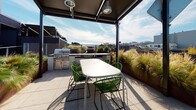 This rooftop terrace offers a well-designed outdoor living experience elevated above the surrounding cityscape. The space is finished with large-format porcelain tile pavers that lend a clean, contemporary aesthetic underfoot, while sleek metal mesh perimeter railings ensure unobstructed sightlines across a broad urban panorama. Generously sized Cor-Ten steel raised planters are filled with ornamental feather grasses and deciduous trees, introducing a natural, textural element that softens the rooftop's modern lines and provides a sense of privacy.

A standout feature is the covered outdoor dining area, sheltered beneath a structured pergola with an integrated overhead canopy. This zone is equipped with a stainless steel built-in BBQ grill and outdoor kitchen setup, complemented by a long dining table and vibrant green chairs — ideal for al fresco entertaining. The pergola provides comfortable shade while remaining open to the surrounding views and sky.

Vertical architectural elements, including ribbed metal cladding in deep navy and warm bronze tones, frame the access points and service structures, adding visual depth and a refined industrial character. The terrace benefits from abundant natural light and open sky exposure, making it a bright and inviting outdoor retreat throughout the day.