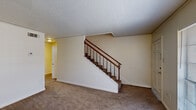 This two-story townhouse in Baton Rouge, Louisiana, offers a practical and straightforward layout distributed across two floors. The ground floor houses the main living area and kitchen, while the upper floor contains the bedroom, bathroom, closet, and hallway, connected by a staircase featuring a classic wood banister with dark-stained spindles.

The living area on the ground floor is carpeted and finished with neutral cream-toned walls and a textured popcorn ceiling. A centrally located staircase anchors the space, leading to the upper level. The kitchen is equipped with dark wood cabinetry, light-toned countertops, a stainless steel sink, tile backsplash, and wood-look flooring, providing a functional cooking and prep area.

Upstairs, the generously sized primary bedroom is carpeted and features a window that allows natural light and outdoor views. Neutral walls and a textured ceiling carry through from the lower level. Adjacent to the bedroom is a dedicated closet space outfitted with built-in shelving and a hanging rod, offering organized storage. The full bathroom includes a tub-shower combination with a curtain rod, a vanity with a sink, tile wainscoting, and a wall-mounted mirror. Throughout, the townhouse maintains a clean, understated aesthetic with consistent neutral finishes.