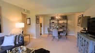 This single-floor residence offers a warm and inviting layout designed for comfortable everyday living. The open-concept living and dining area serves as the heart of the home, bathed in soft, ambient light that highlights the neutral-toned walls and plush carpeted flooring throughout. The dining area is a standout feature, anchored by a round table surrounded by upholstered chairs and accented by a striking industrial-style orb chandelier overhead. A stylish open shelving unit provides both display space and decorative charm, adding character to the room. The living area flows naturally from the dining space, offering a cozy setting furnished with complementary accent pieces, including a mirrored side table and tasteful wall art that contribute to the refined aesthetic. A wide, open passageway connects the main living area to the kitchen, which is visible in the background and features light cabinetry, providing easy accessibility and a sense of continuity throughout the home. Overhead HVAC vents are integrated into the ceiling, ensuring year-round comfort. The overall ambiance is polished yet approachable, blending contemporary decorative elements with a classic residential layout that feels both functional and welcoming.