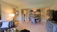 This single-floor residence offers a warm and inviting layout designed for comfortable everyday living. The open-concept living and dining area serves as the heart of the home, bathed in soft, ambient light that highlights the neutral-toned walls and plush carpeted flooring throughout. The dining area is a standout feature, anchored by a round table surrounded by upholstered chairs and accented by a striking industrial-style orb chandelier overhead. A stylish open shelving unit provides both display space and decorative charm, adding character to the room. The living area flows naturally from the dining space, offering a cozy setting furnished with complementary accent pieces, including a mirrored side table and tasteful wall art that contribute to the refined aesthetic. A wide, open passageway connects the main living area to the kitchen, which is visible in the background and features light cabinetry, providing easy accessibility and a sense of continuity throughout the home. Overhead HVAC vents are integrated into the ceiling, ensuring year-round comfort. The overall ambiance is polished yet approachable, blending contemporary decorative elements with a classic residential layout that feels both functional and welcoming.