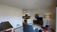 This single-floor unit offers an open-concept layout that seamlessly connects the living area and kitchen, creating an efficient and inviting space. The kitchen is outfitted with white cabinetry, stainless steel appliances including a range and over-the-range microwave, and a dark tile backsplash that adds visual contrast against the clean white surfaces. The living area is styled with mid-century modern influences, featuring a blue accent chair, a wooden media console with tapered legs, and a flat-screen television. A bold abstract artwork in mustard yellow, gray, and blue tones anchors the living space, adding a pop of color to the neutral white walls. The flooring transitions between hardwood and a geometric-patterned area rug that defines the seating zone. Recessed lighting and flush-mounted fixtures provide consistent illumination throughout the space. A floor-level vent and baseboard details are visible, reflecting standard residential construction. The overall ambiance is modern yet comfortable, with a curated aesthetic that balances functionality and style.