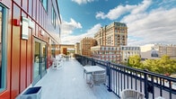 Located at 1112 First St in the heart of Old Town Alexandria, Virginia, this rooftop terrace offers an impressive outdoor experience defined by sweeping panoramic views of the surrounding Alexandria cityscape. The terrace stretches across a generous elongated deck finished with light-toned composite wood planking, providing a clean, modern aesthetic underfoot.

The space is bordered by a sleek navy blue metal railing system that frames unobstructed views of Alexandria's historic and contemporary architecture, including prominent brick mid-rise buildings that define the city's skyline. Lush tree canopies and open sky complete the scenic backdrop, making this an ideal setting for outdoor gatherings.

A bold, vertical red metal panel facade lines the building exterior adjacent to the terrace, lending a striking visual contrast to the light deck surface and blue railings. Wall-mounted cylindrical light fixtures are integrated into the facade, providing functional illumination for evening use. Large sliding glass doors connect the terrace to the interior, allowing for seamless indoor-outdoor flow.

Seating throughout the terrace is thoughtfully arranged in multiple zones, featuring Adirondack chairs, woven lounge chairs, side tables, and dining-height table-and-chair sets — accommodating both relaxed lounging and alfresco dining. Planter containers positioned along the deck add greenery to the environment. The terrace receives abundant natural light, making it a vibrant and inviting amenity space throughout the day.