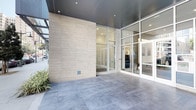 This contemporary single-floor space makes a strong first impression with its thoughtfully designed exterior entrance. Large floor-to-ceiling glass panels and aluminum-framed storefronts invite natural light deep into the interior, offering clear visibility and an open, welcoming atmosphere. The facade combines warm, light-toned wood panel cladding with a dark metallic ceiling soffit, creating a refined contrast that speaks to modern architectural sensibility. Underfoot, large-format grey porcelain tiles extend across the entry area, lending a clean and polished aesthetic that carries through the space. Recessed lighting is strategically placed throughout, ensuring consistent, well-distributed illumination that complements the natural light. Inside, the open layout is visible through the glazed entry doors, hinting at a bright and organized interior with white walls and a neutral palette that enhances the sense of openness. Touches of greenery near the entrance add a subtle organic element to the otherwise structured, minimalist design. The overall ambiance is professional yet approachable, making this space well-suited for a variety of commercial uses. The clean lines, quality finishes, and contemporary detailing come together to create a polished and functional environment.